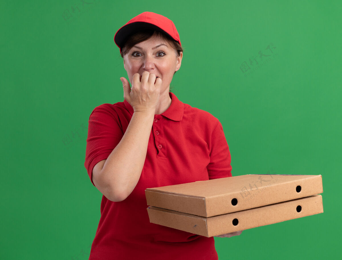 制服身穿红色制服 头戴鸭舌帽的中年女送货员拿着披萨盒 看着站在绿色墙壁上紧张不安 咬人的指甲中年女人盒子