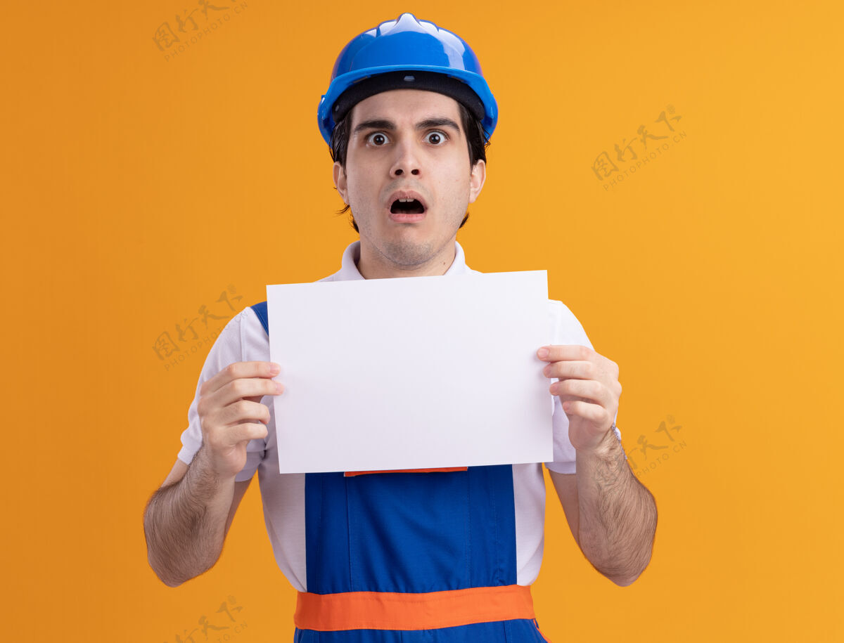 担心年轻的建筑工人穿着建筑制服 戴着安全帽 手里拿着空白的一页 站在橘色的墙上 惊讶又担心地看着前面年轻头盔惊人