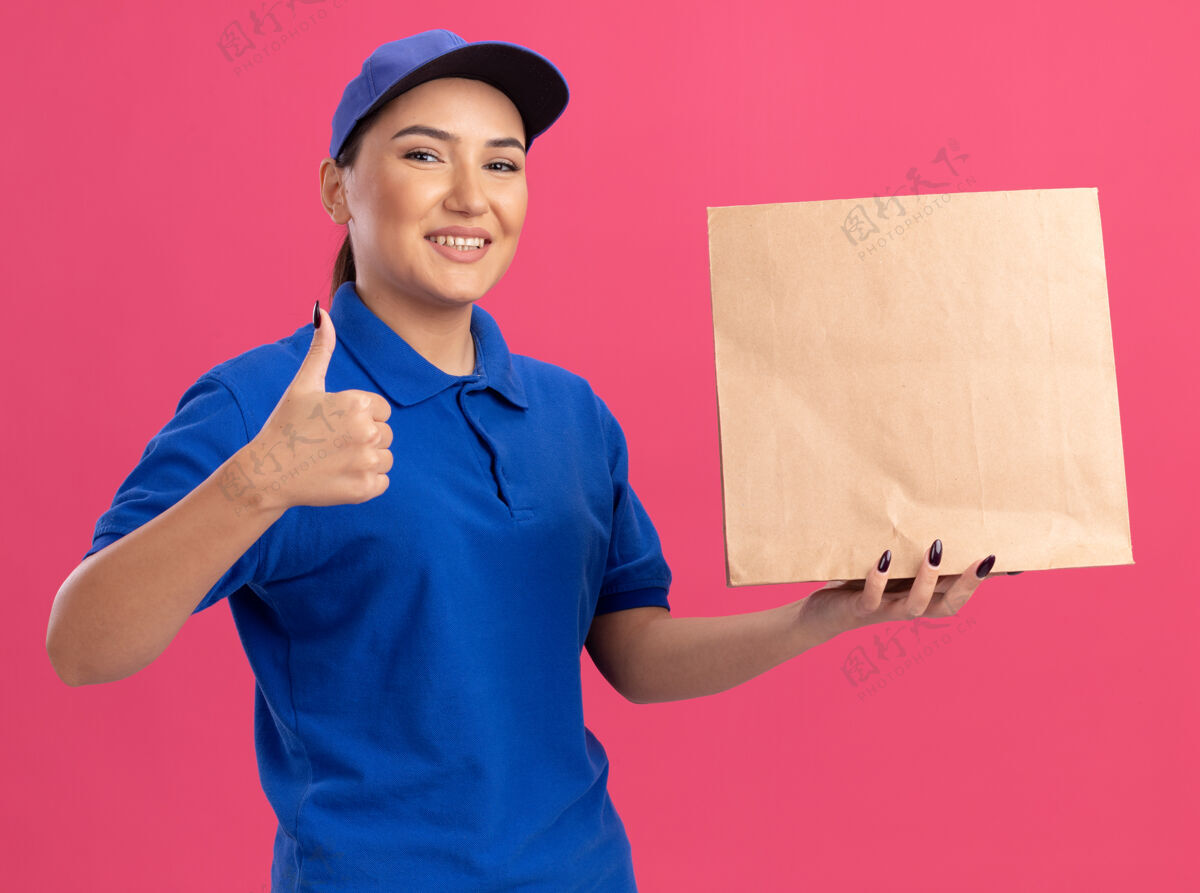 微笑身着蓝色制服 头戴鸭舌帽 拿着纸包的快乐女送货员站在粉红色的墙上 面带微笑 高高兴兴地竖起大拇指站着纸年轻