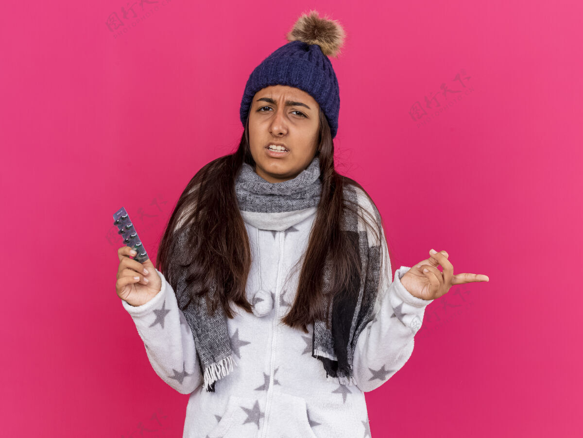 年轻困惑的年轻生病的女孩戴着冬天的帽子和围巾拿着药片和点在一边孤立的粉红色背景与复制空间围巾抱壁板