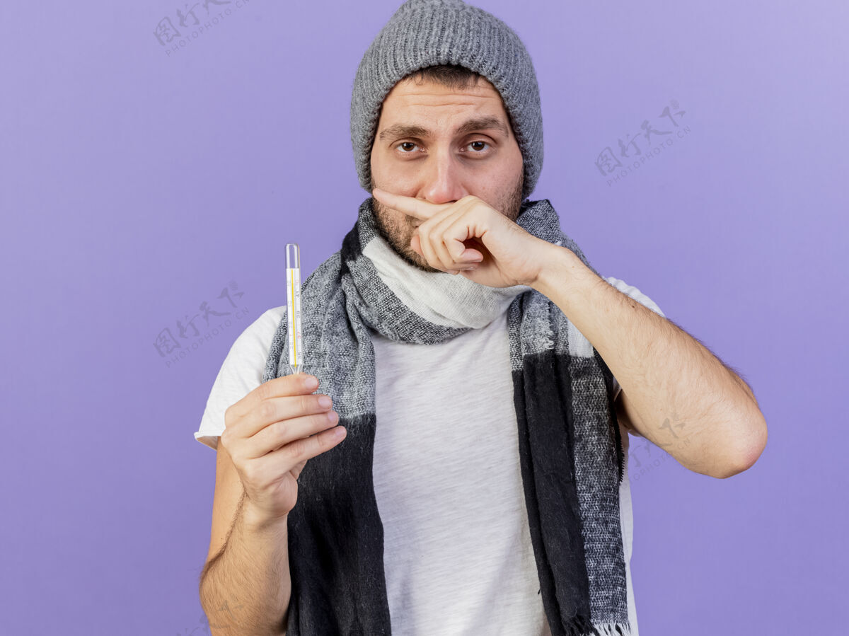 穿未出狱的年轻病人戴着冬天的帽子 戴着围巾 拿着温度计 用手在紫色背景上擦鼻子手湿巾围巾