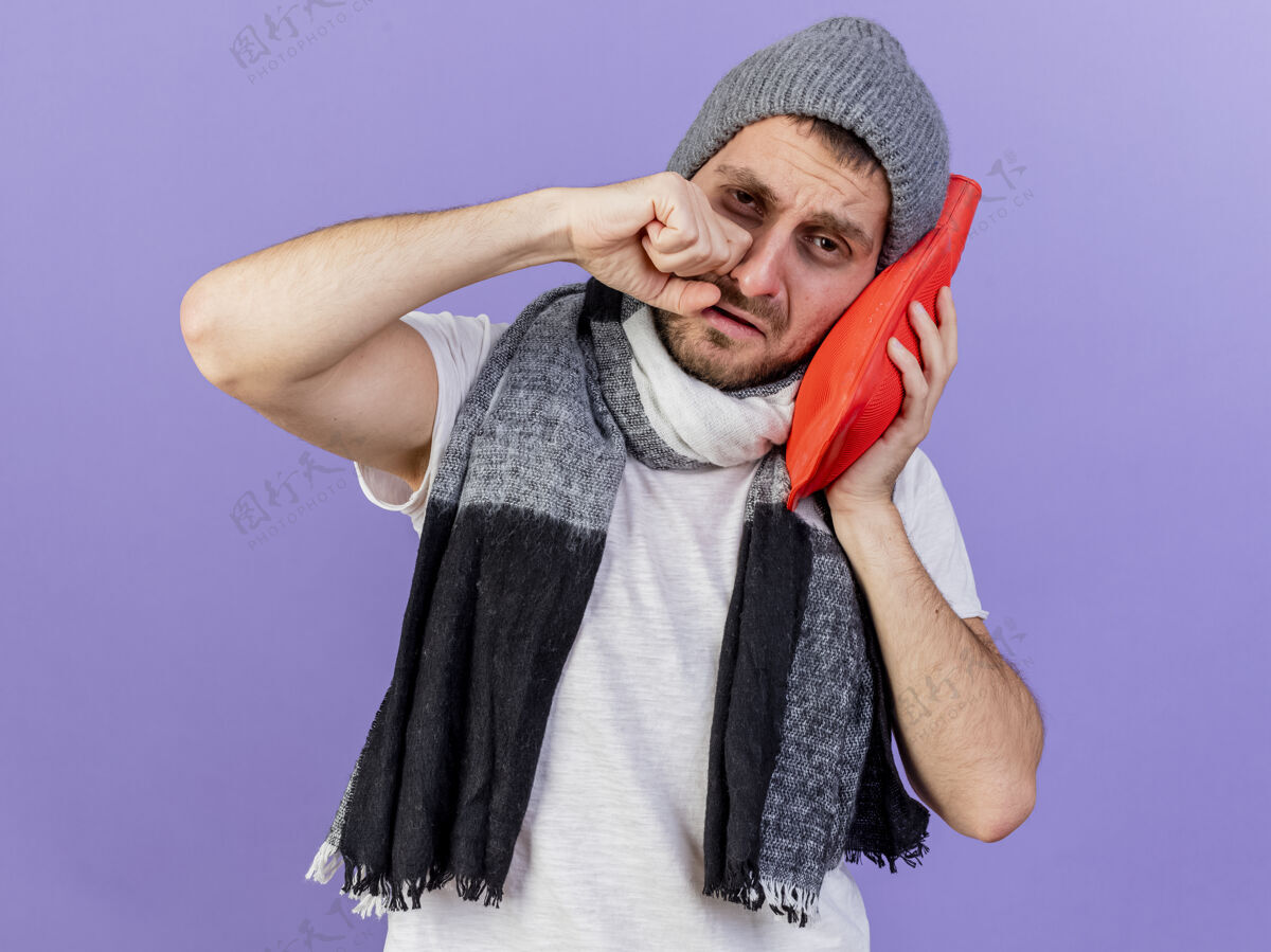 帽子哭泣的生病的年轻人戴着冬天的帽子 戴着围巾 脸颊上拿着热水瓶 在紫色的背景下与世隔绝抱病年轻人