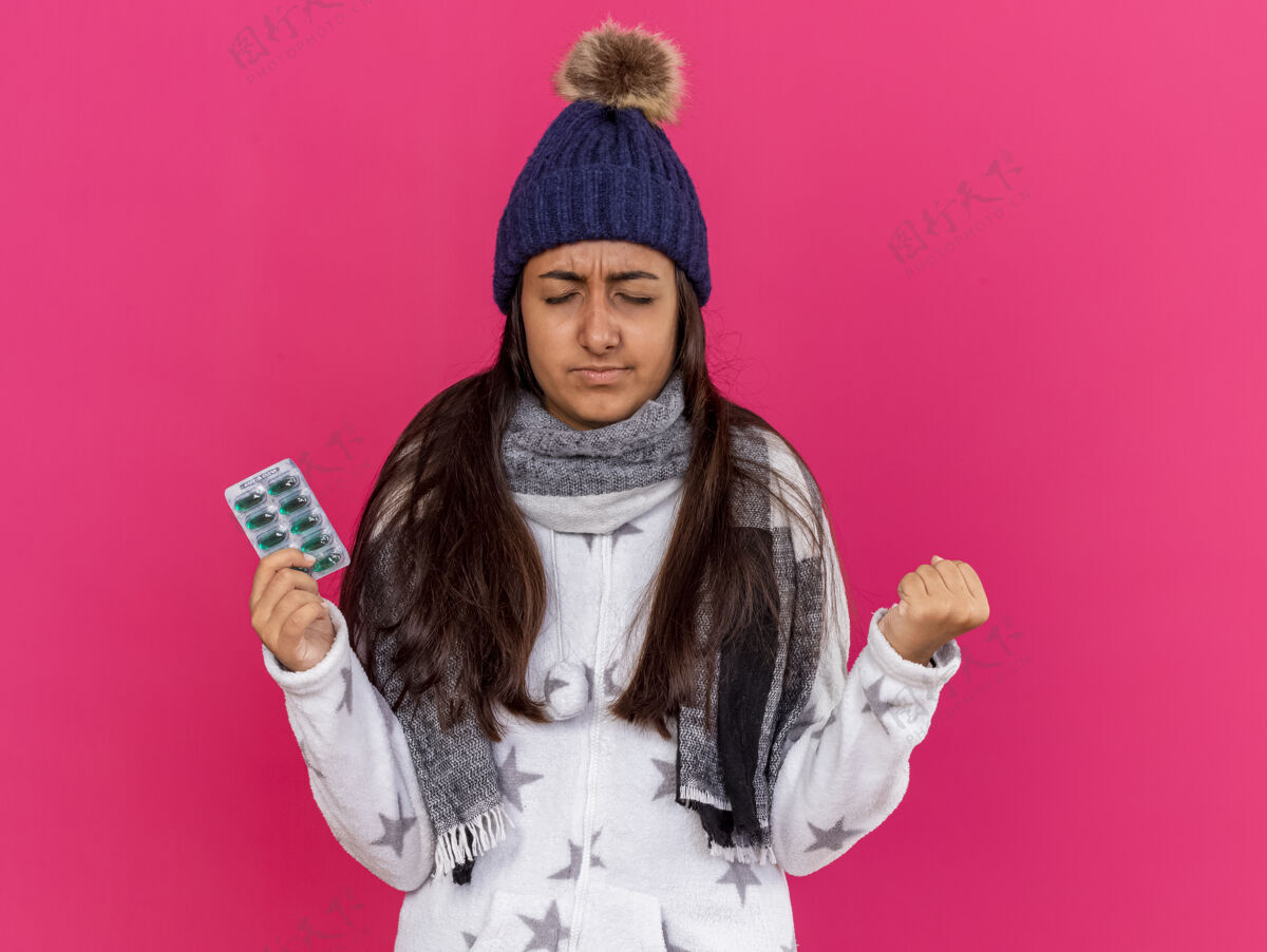 抱着关心的年轻病女孩闭着眼睛戴着冬天的帽子围巾拿着隔离在粉红色背景上的药丸戴着关心药片