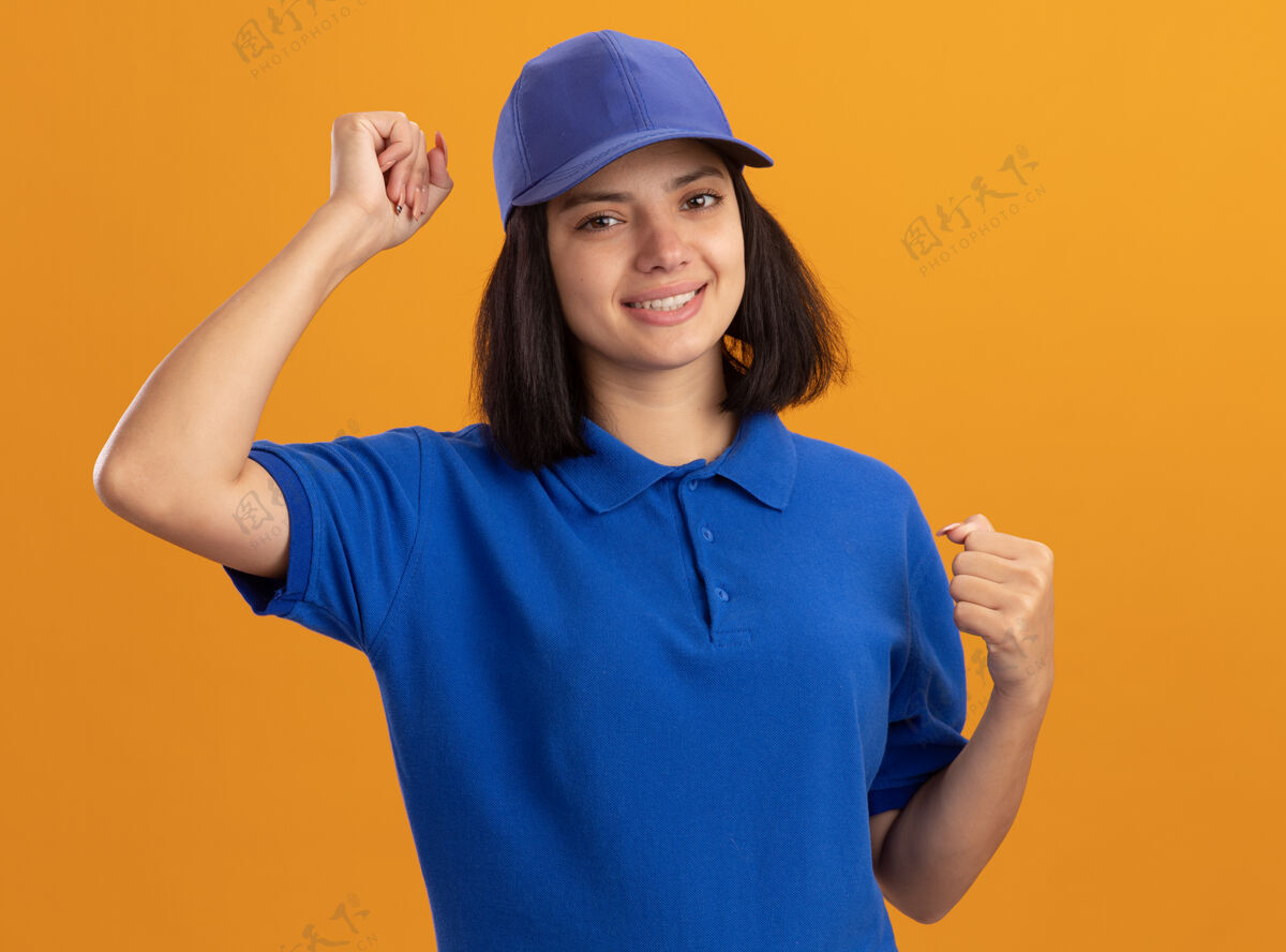 帽子身穿蓝色制服 头戴鸭舌帽的年轻送货员高兴而兴奋地握紧拳头 为自己站在橘色的墙上的成功而欢欣鼓舞成功女孩拳头