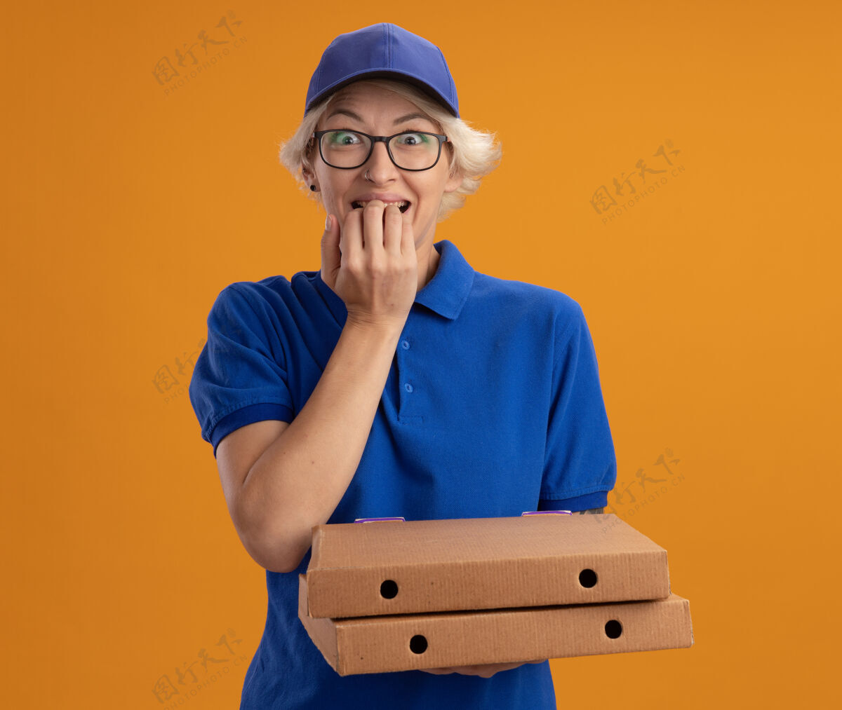 咬身穿蓝色制服 戴着眼镜 拿着比萨饼盒的年轻送货员紧张地咬着橘色墙上的钉子拿着紧张盒子