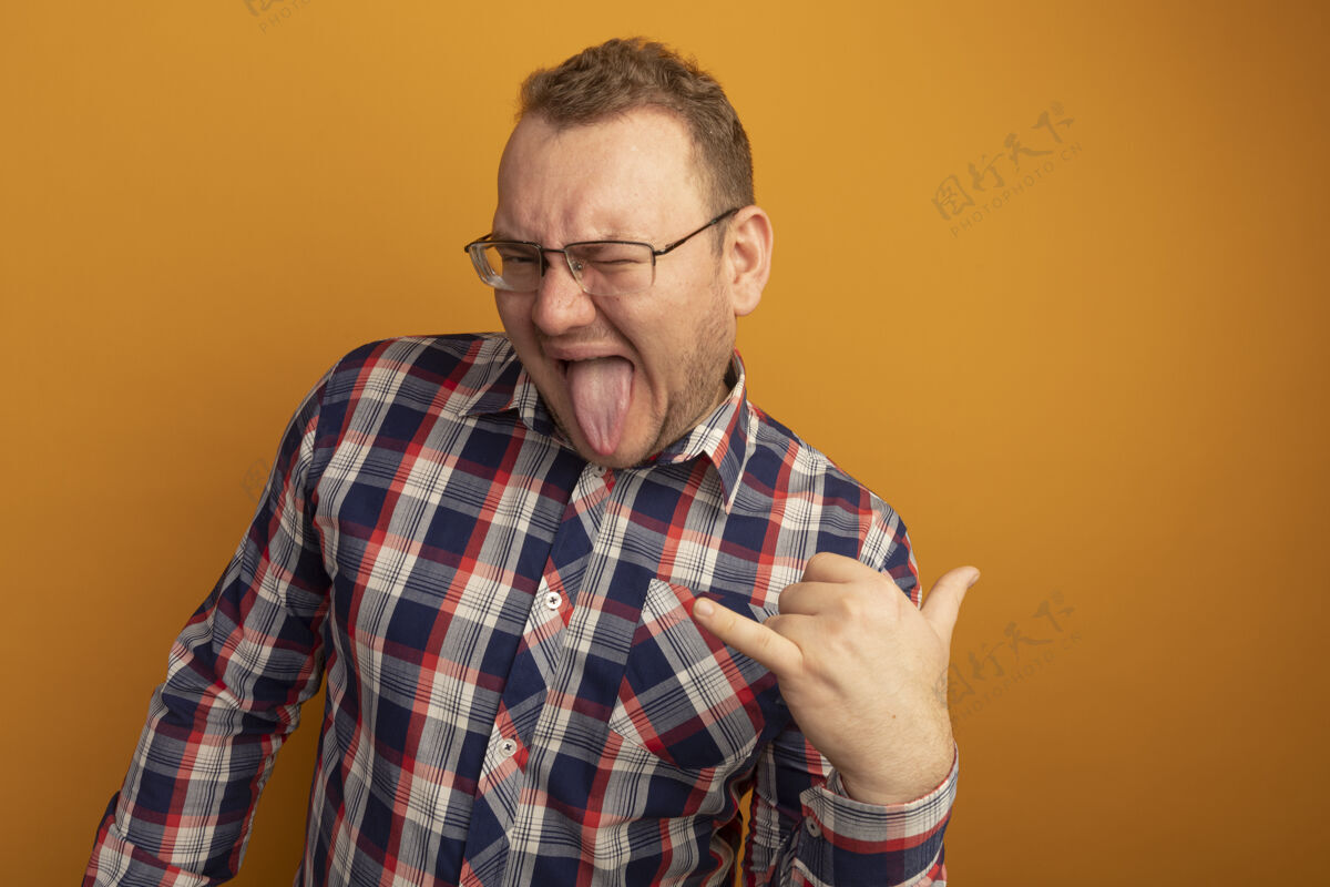坚持戴着眼镜 穿着格子衬衫的快乐男人快乐快乐地展示着站在橙色墙壁上伸出舌头的岩石符号欢呼站立男人