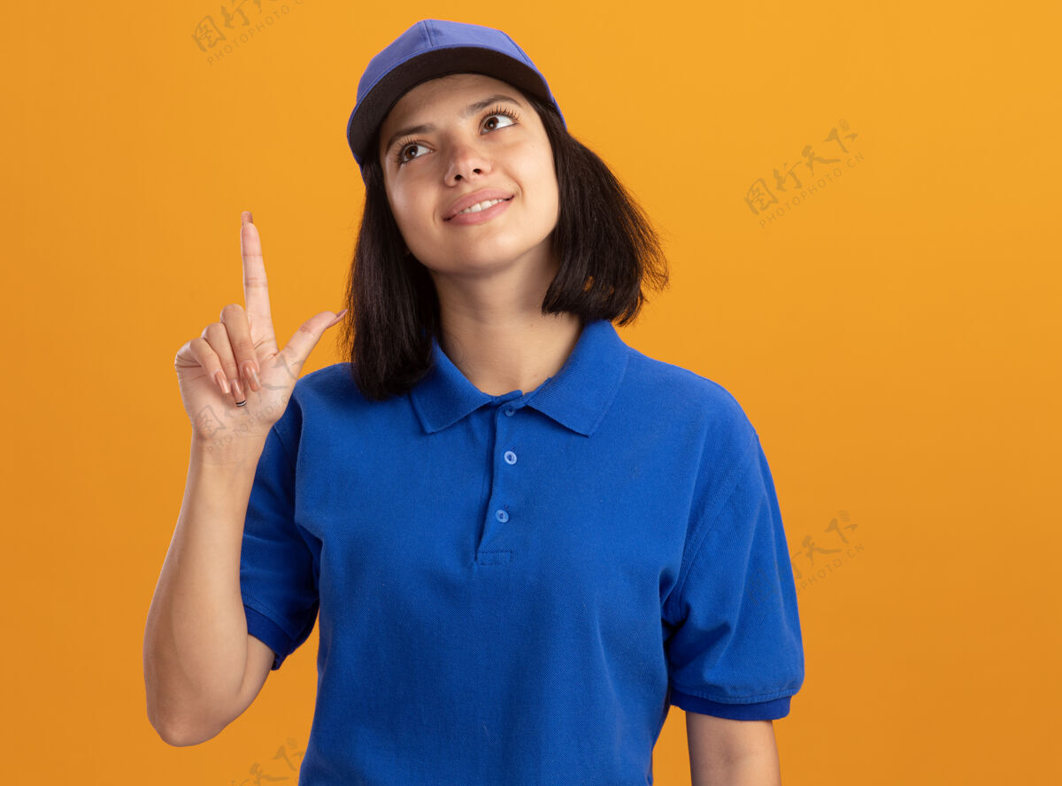 指着身穿蓝色制服 戴着帽子 站在橙色的墙上微笑着向上指指点点的年轻女送货员目录女孩年轻人