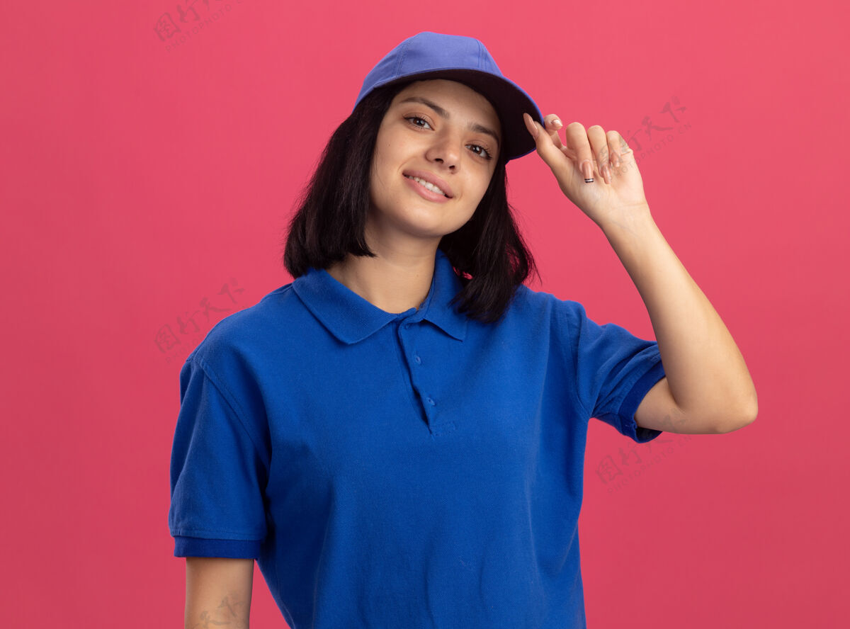 帽子身穿蓝色制服 戴着帽子的年轻送货女孩站在粉红色的墙上 自信地微笑着修好帽子信心微笑修复
