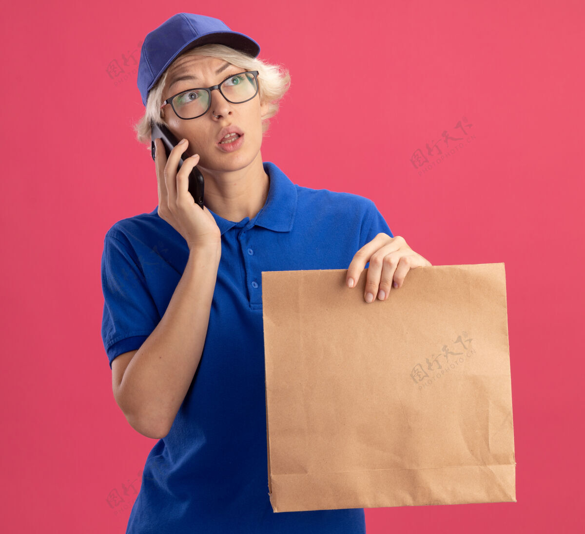 包装穿着蓝色制服 戴着帽子的年轻送货员拿着纸包在手机上聊天 隔着粉色的墙 一边困惑地看着一边女人年轻人帽子