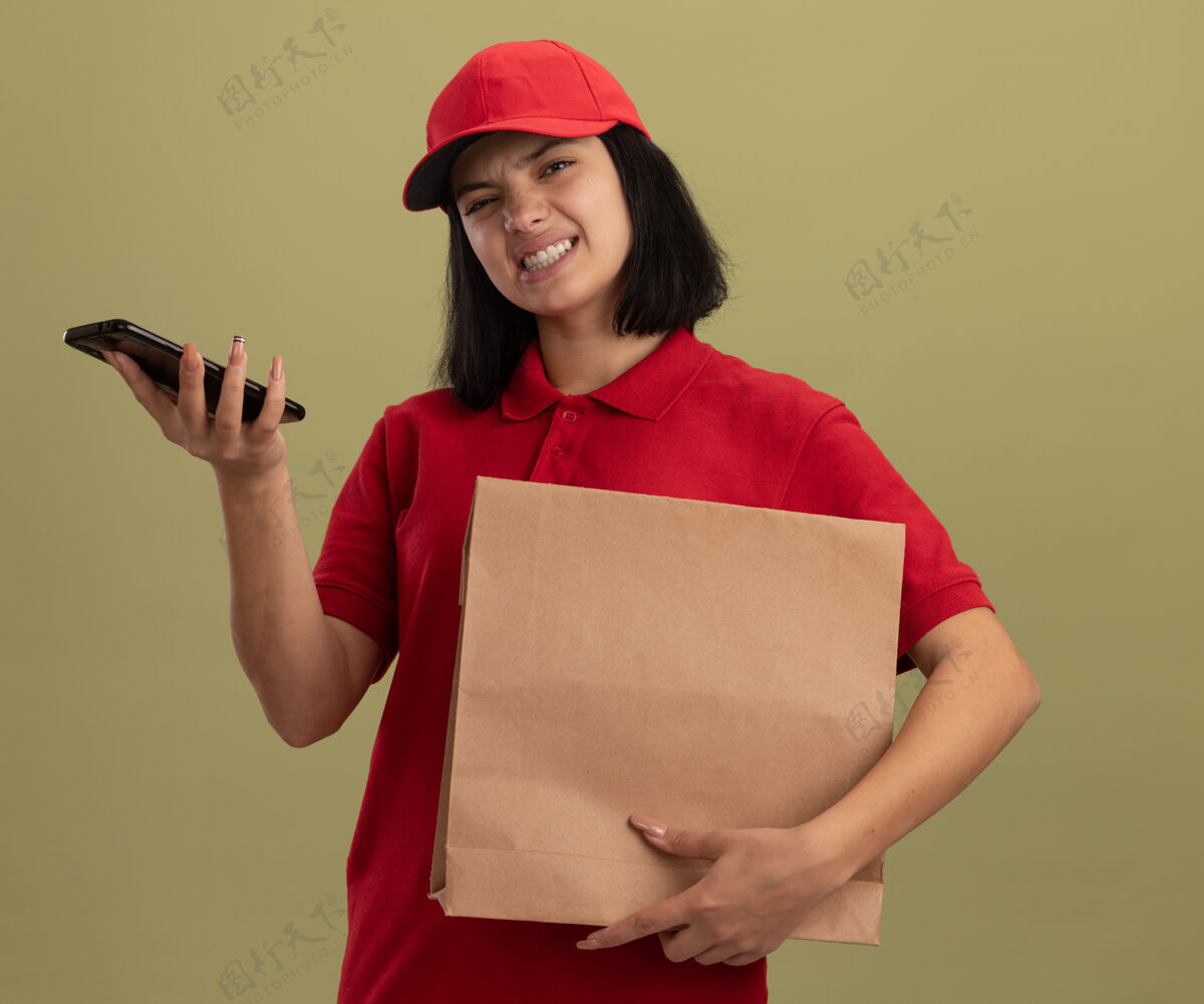 包装穿着红色制服 戴着帽子 拿着纸包的年轻送货女孩站在明亮的墙上 脸上带着恼怒的表情交货立场恼火