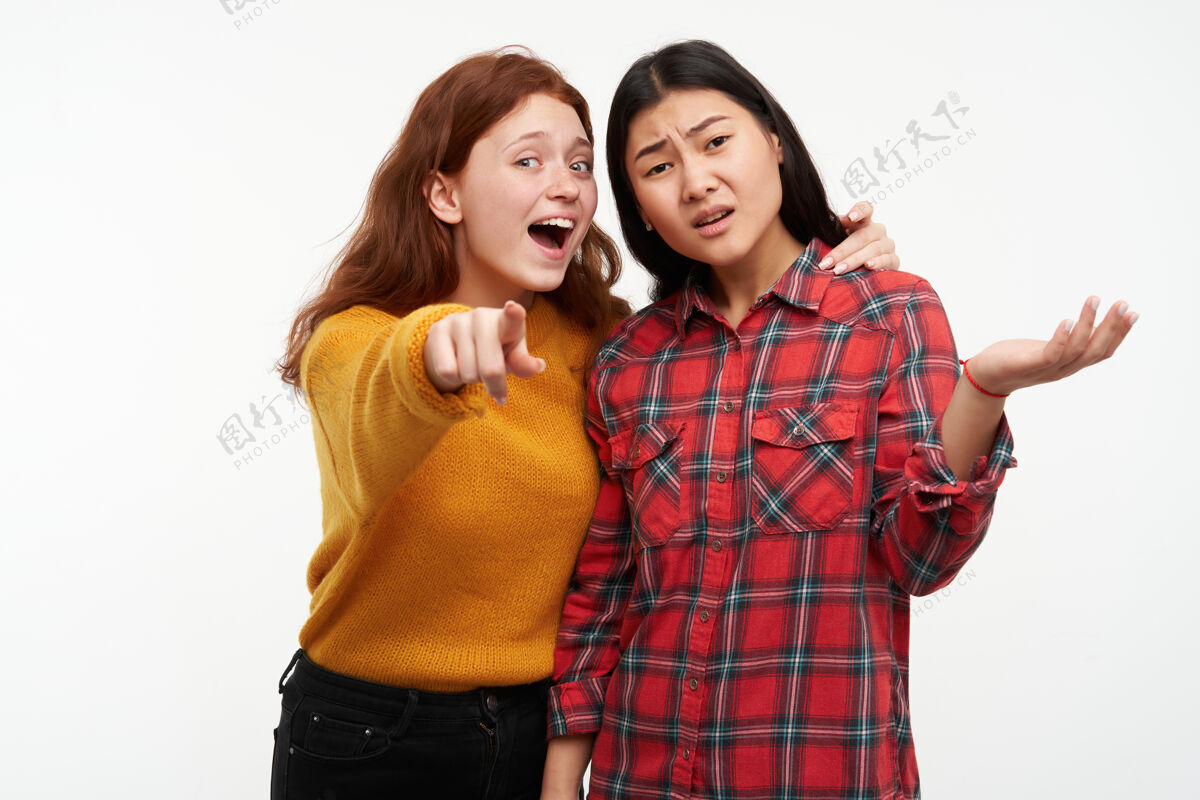 指向两个年轻的女性朋友一个女孩给她的朋友看东西 但另一个看不见穿着黄色毛衣和格子衬衫隔着白色的墙表演青年朋友