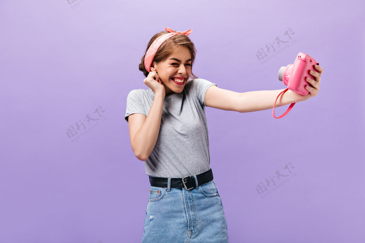 成人快乐的女人拿着粉红色的相机自拍快乐迷人的女士穿着灰色t恤和牛仔裙 配上黑色腰带拍照头带表情紫色
