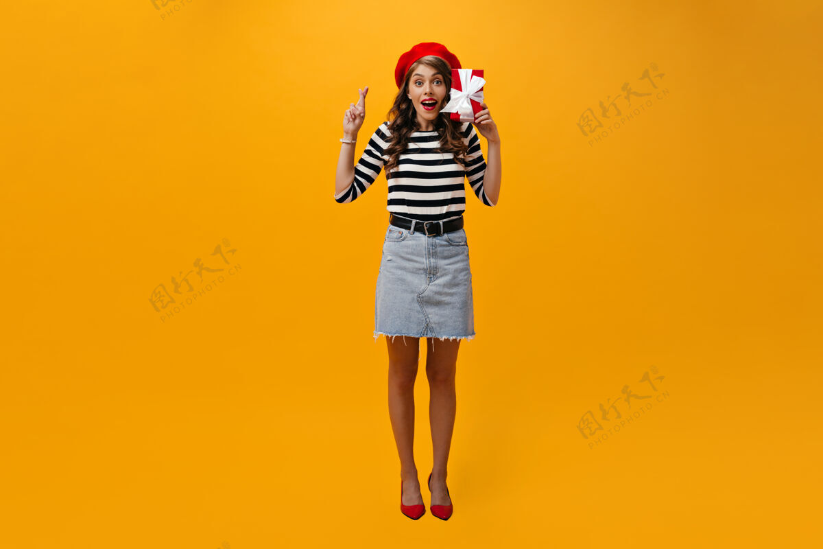 橙色女人交叉手指 拿着红色礼盒的全长镜头穿着红色贝雷帽和条纹衬衫的快乐女孩看着镜头微笑牛仔裙女士