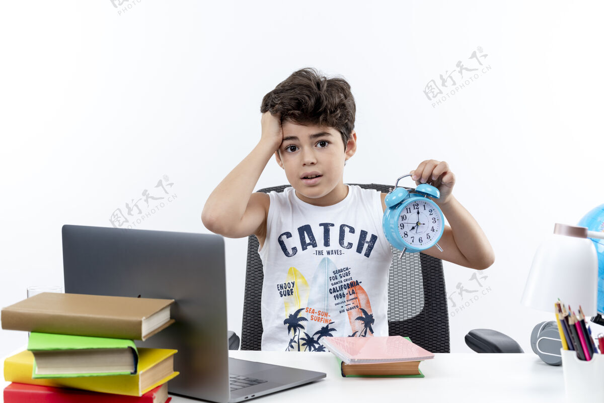 学校关心的小男孩坐在书桌旁 手里拿着学习工具 手里拿着闹钟 抓起孤立在白色背景上的头小抓住工具