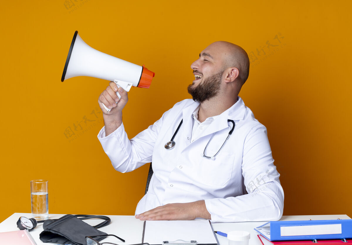 说话看着旁边快乐的年轻秃头男医生穿着医用长袍和听诊器坐在办公桌上 拿着医疗工具 用橙色背景隔离的扬声器说话穿着壁板工具