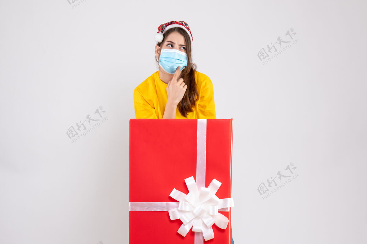 礼物前视图好奇的圣诞女孩站在圣诞礼物后面 戴着圣诞帽丝带节日大