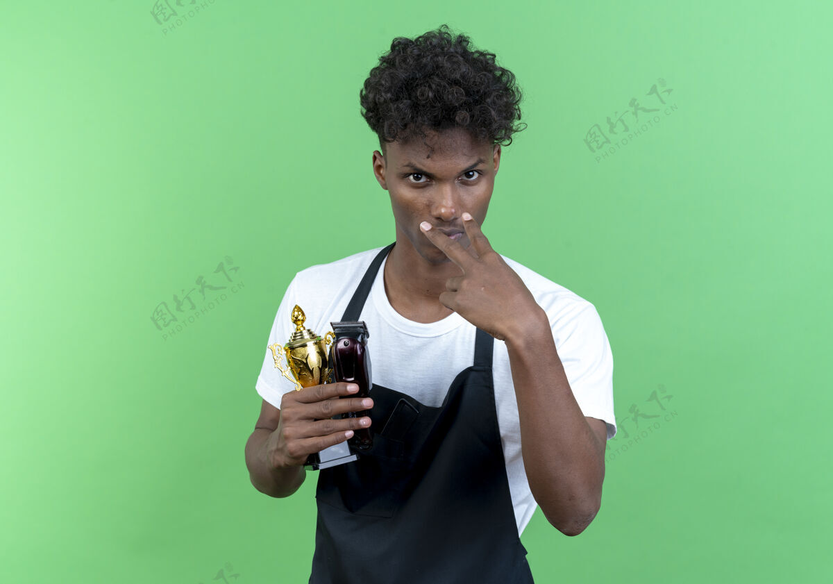 观看看着镜头年轻的黑人男理发师穿着制服拿着优胜者杯 在绿色的背景下看你的手势孤立年轻人男性优胜者