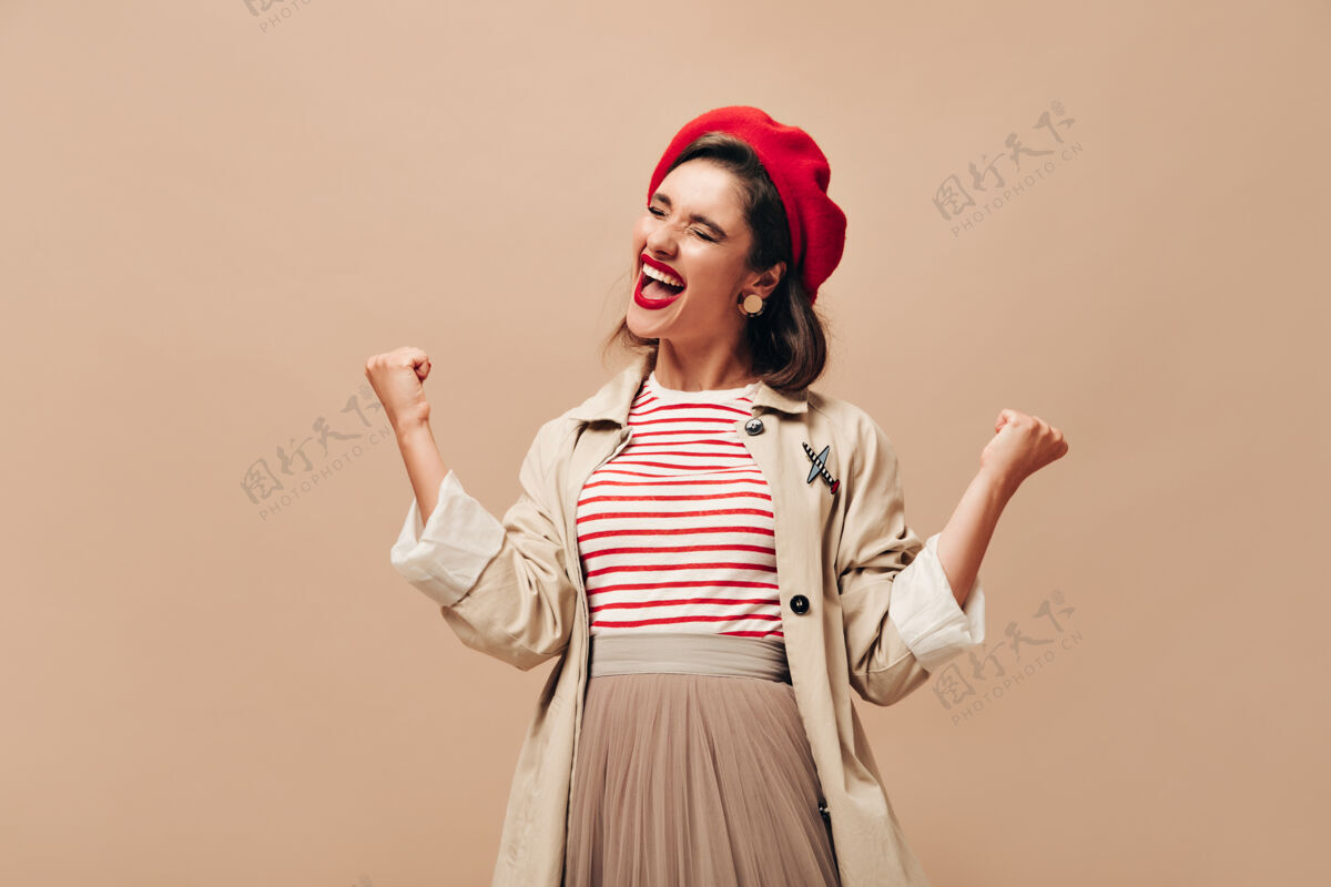 休闲穿着时髦风衣戴着帽子的女士在米色背景下情绪化地摆姿势戴着红色贝雷帽的黑发快乐的年轻女子欢欣鼓舞女性站立裙子