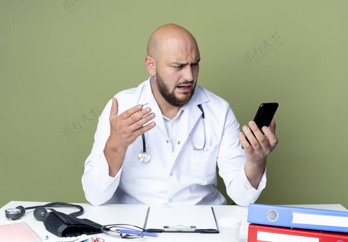 坐着愤怒的年轻秃头男医生穿着医用长袍和听诊器坐在办公桌前 手持医疗工具 孤立地看着绿色背景上的电话办公桌男性穿