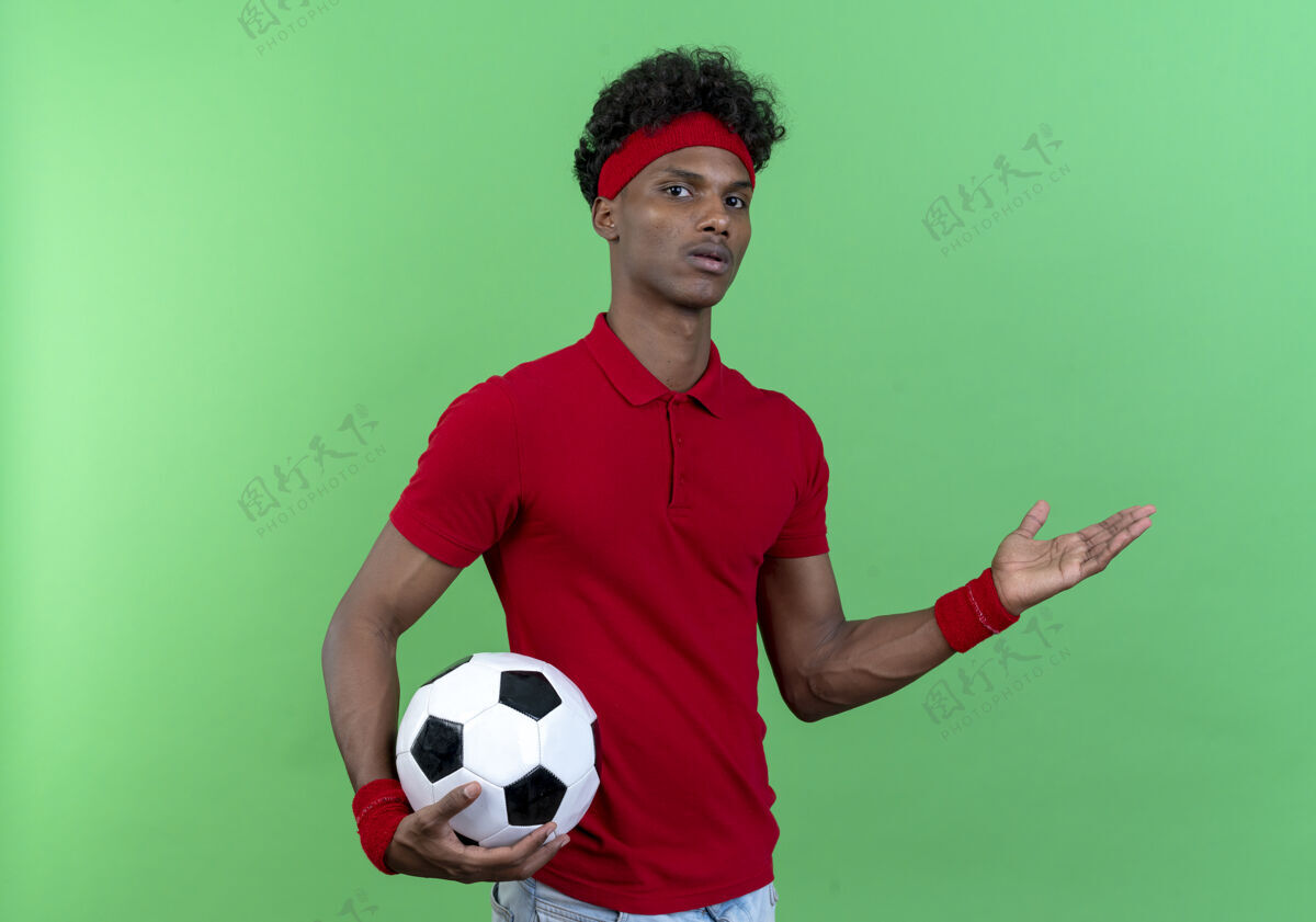 壁板困惑的年轻美国黑人运动男子戴着头带和手环拿着球和点与手侧隔离在绿色背景与复制空间年轻穿运动