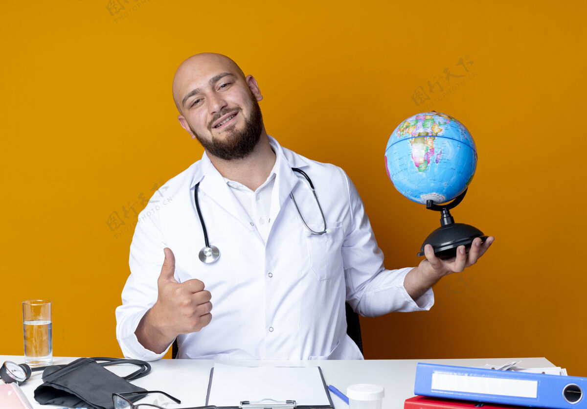 听诊器年轻的秃顶男医生穿着医用长袍和听诊器坐在办公桌旁 手里拿着医疗工具 把拇指孤立地举在橙色的背景上秃头请穿办公桌