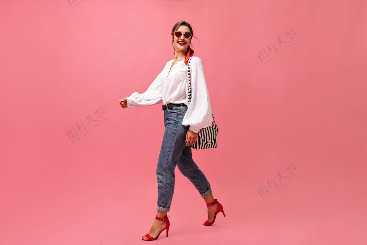 时尚穿着牛仔裤 白衬衫 在粉色背景上行走的微笑女士一个戴着红色太阳镜的时髦女人走在孤立的背景上姿势时尚空白空间