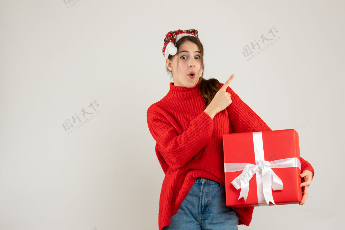 抱着前视图好奇的女孩戴着圣诞帽拿着礼物手指指着站着的东西指着微笑圣诞老人