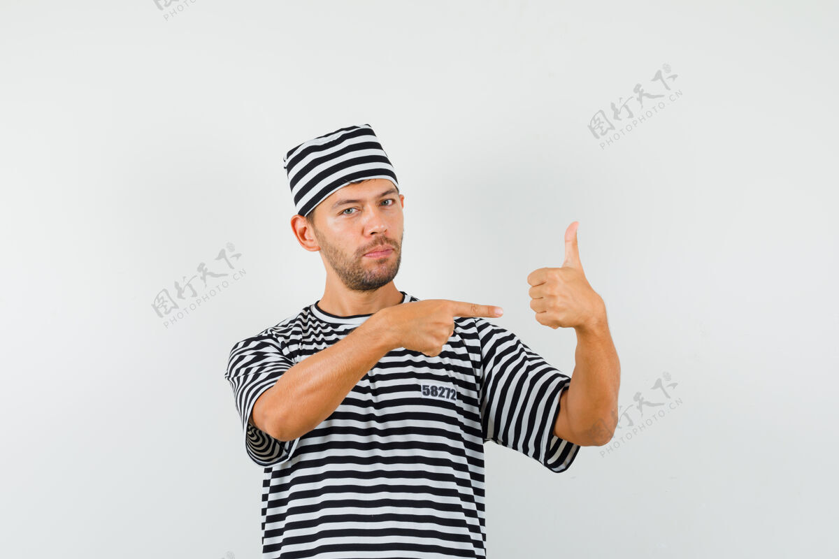 人类戴着条纹t恤帽的年轻男子指着大拇指 看上去很自信胡须商务摄像头表情