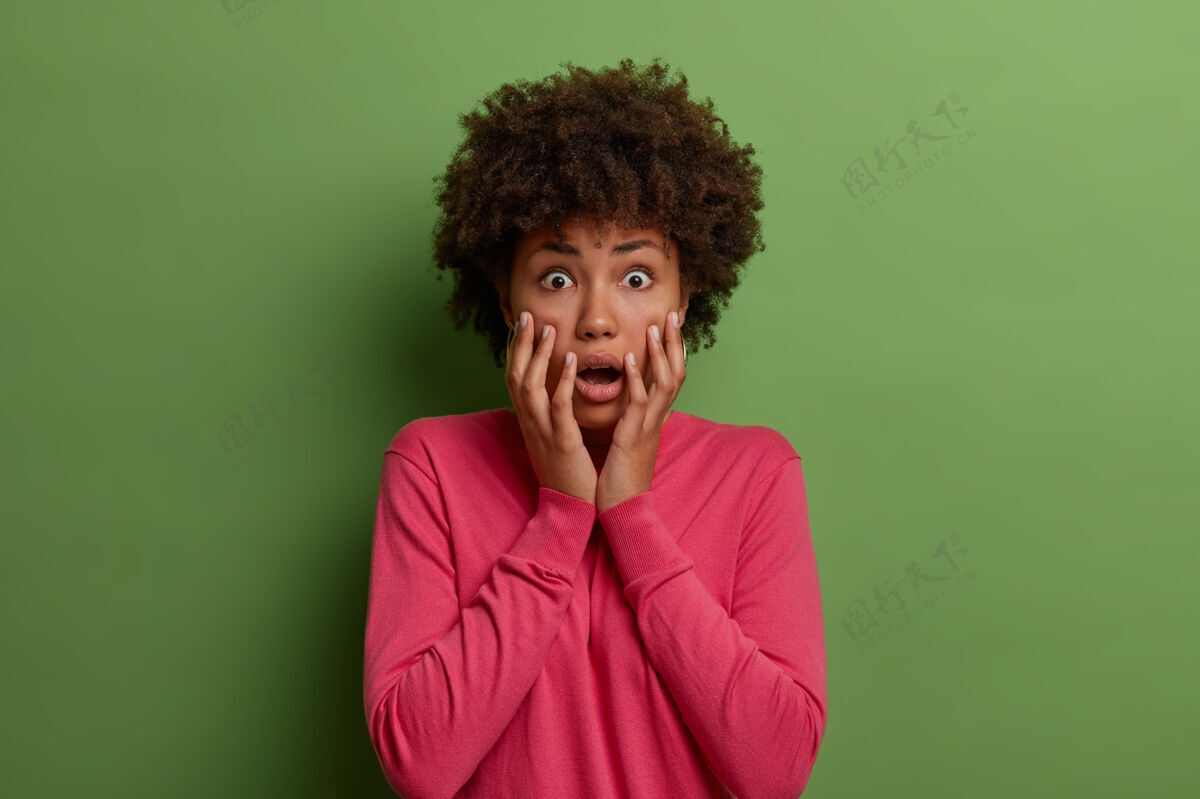 情感没有安全感的惊慌失措的非洲裔美国妇女盯着焦虑 感到震惊和尴尬 手掌放在脸上 穿着粉色的套头衫 意识到绿色墙上有什么令人震惊的孤立的东西肖像生动惊奇