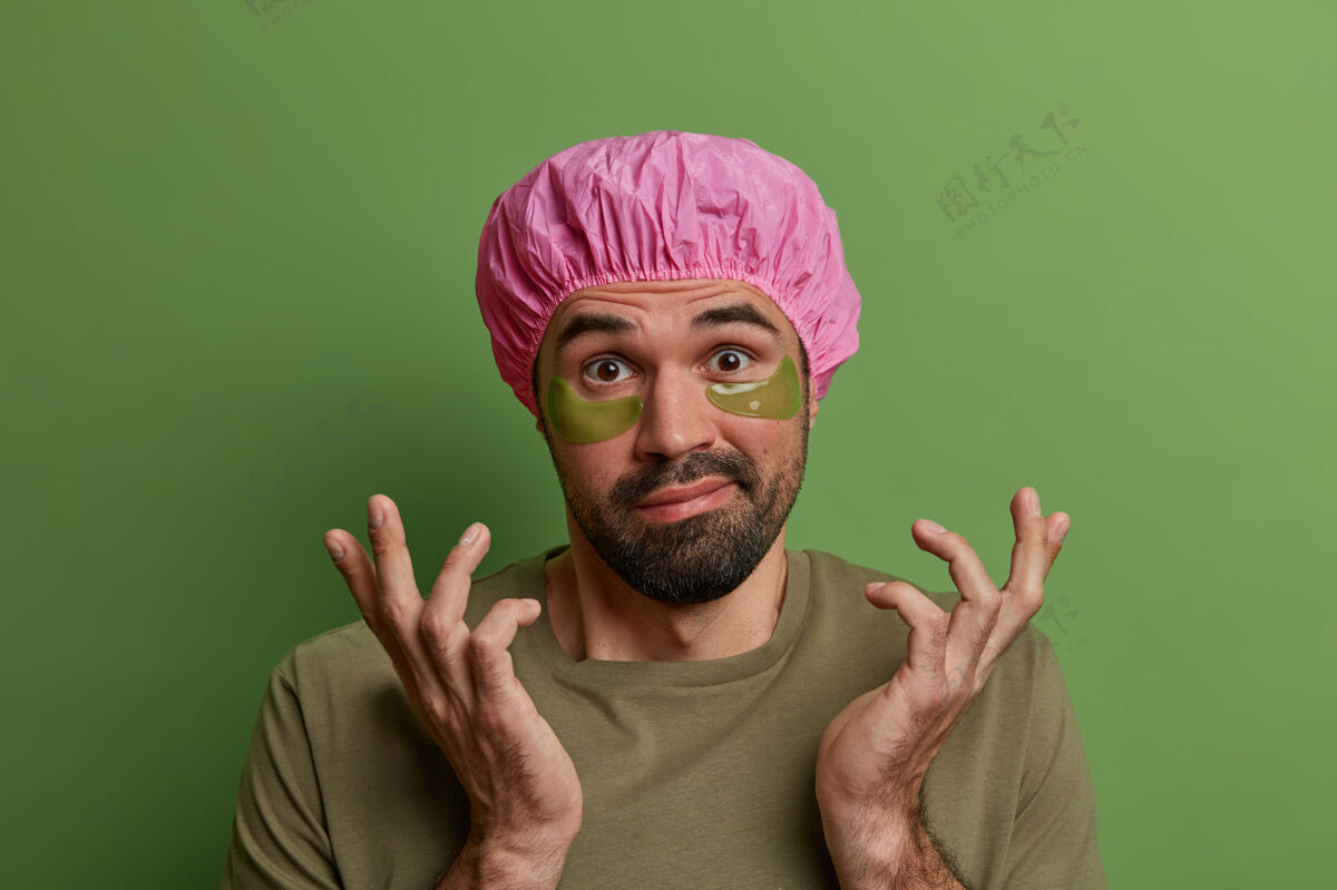 提升个人护理 男士卫生 美容常规概念犹豫不决的欧洲男士将手掌向侧面展开 在眼睛下面贴上眼罩 抚平皱纹 戴上浴帽 靠着绿墙站着圆形面部头部
