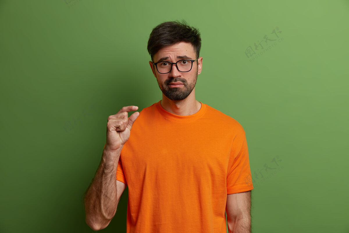 帅气身体语言和尺寸概念年轻人不高兴地展示小尺度 讲述他的微薄工资 塑造一些小东西 判断质量差 穿着橙色t恤 孤立在绿色墙上形状深色橙色