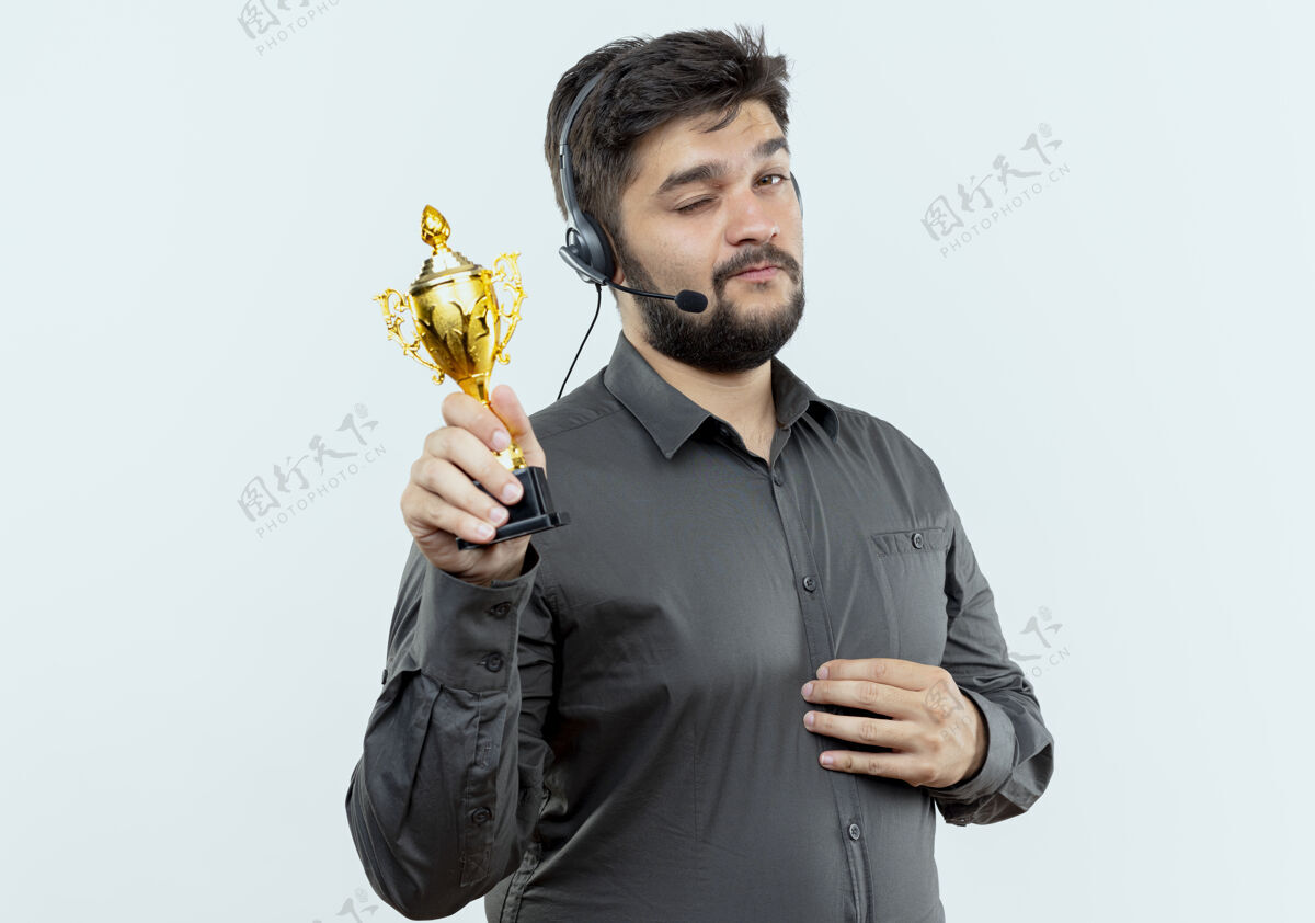 年轻戴着耳机的年轻呼叫中心男子拿着白色背景上孤立的优胜者杯耳机居中手持