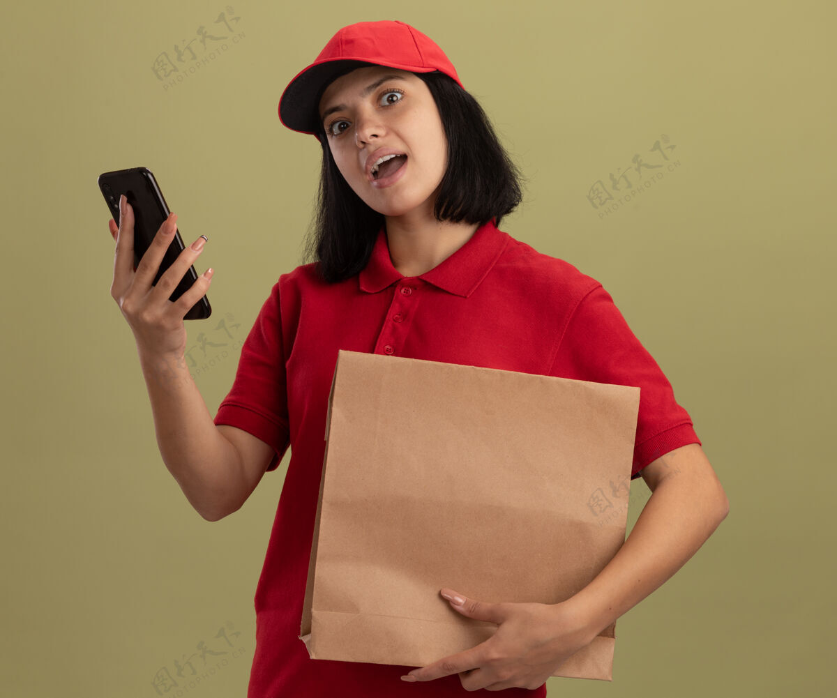 移动年轻的送货员身穿红色制服 戴着帽子 手里拿着纸包 手里拿着智能手机 站在明亮的墙壁上惊喜帽子包装