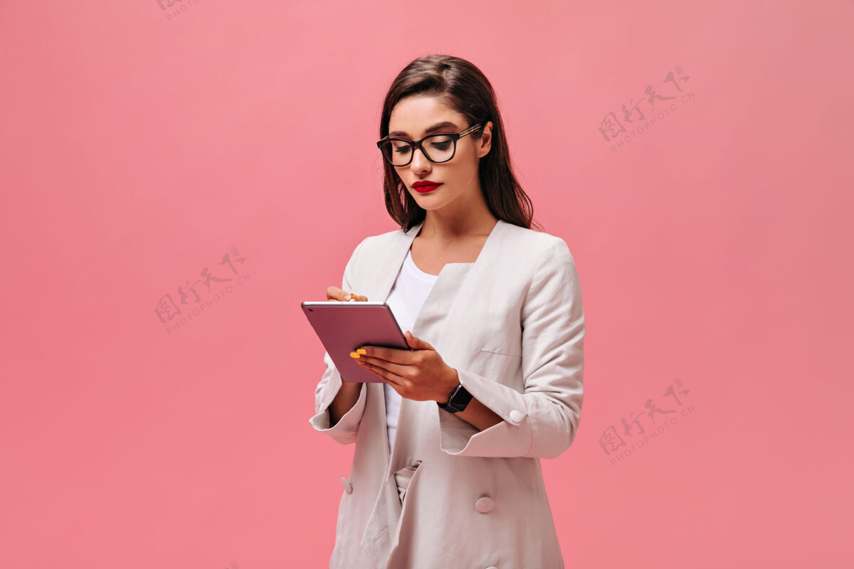高跟鞋戴着眼镜和夹克的女士拿着粉色背景的平板电脑穿着鲜艳衣服的红唇女商人写了些东西年轻平板卷发