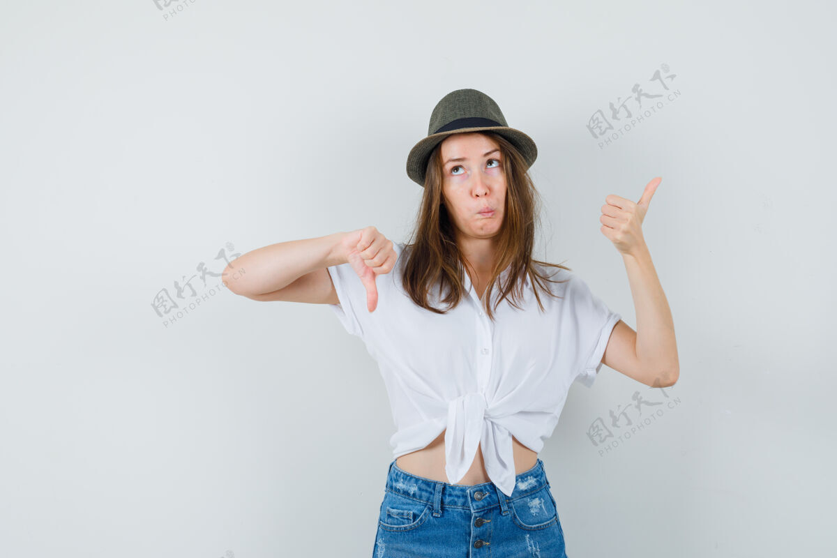 女士穿着t恤牛仔裤帽子上下竖起大拇指的年轻女士 看上去很犹豫显示一起拇指