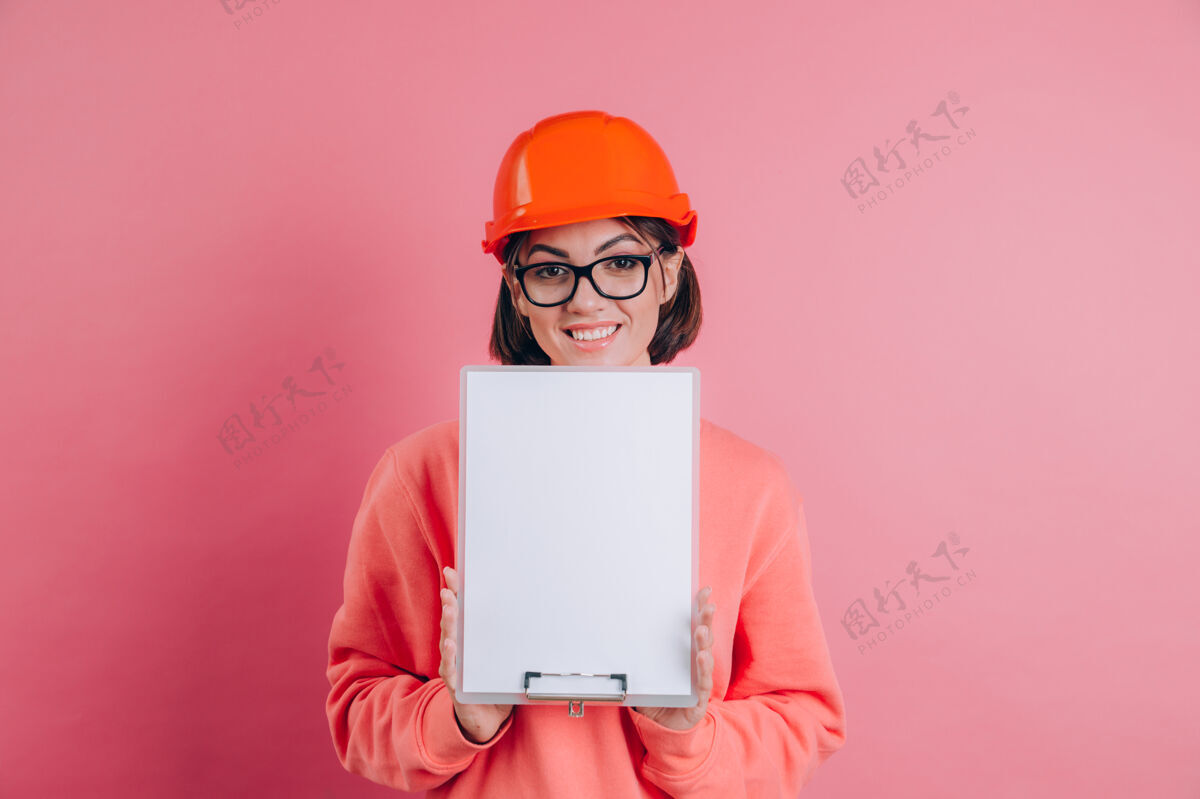 承包商面带微笑的女建筑工人手持白色的空白标志牌 背景是粉色的建筑头盔剪贴板一个帽子