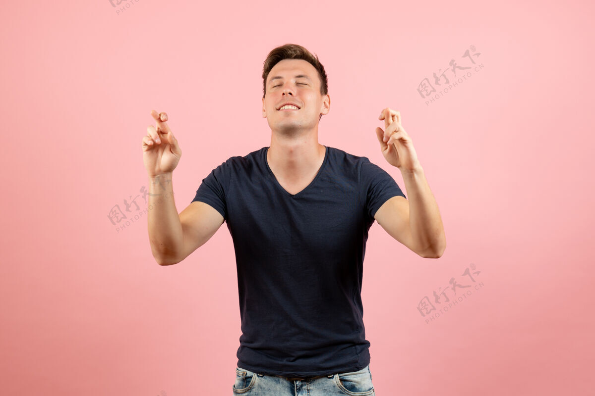 T恤前视图身穿蓝色t恤的年轻男性闭着眼睛交叉着手指 粉色背景上是男性情感色彩的模特人类衬衫封闭眼睛