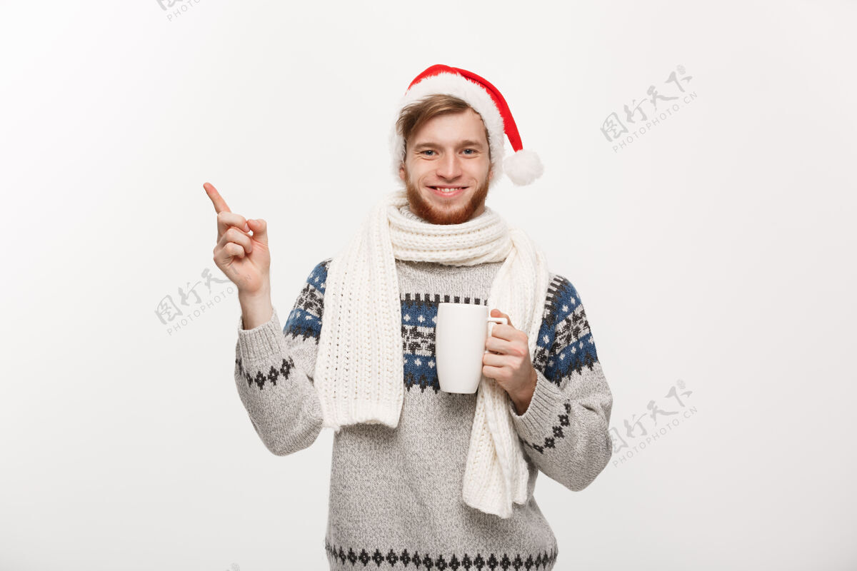 肖像穿着毛衣 戴着圣诞老人帽的年轻胡须男拿着一个热咖啡杯 手放在白色的一边杯子圣诞老人红色