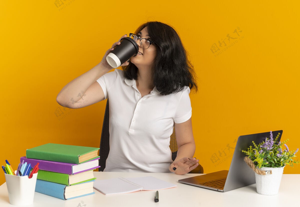 向上年轻快乐漂亮的白人女学生戴着眼镜坐在书桌旁 拿着学习用具喝着咖啡 看着橘黄色和复印空间工具穿咖啡