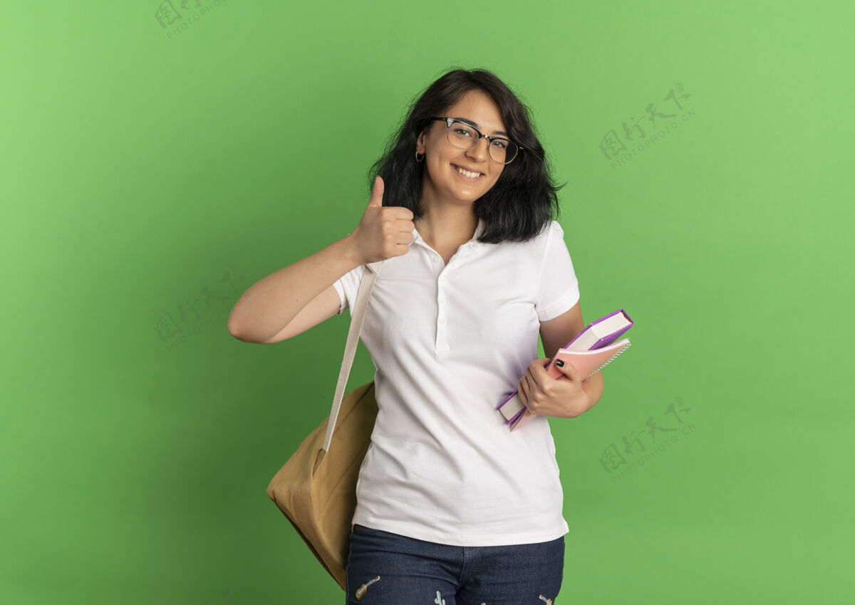 眼镜年轻的高加索女孩 面带微笑 戴着眼镜 背着书包 竖起大拇指 手里拿着一本书 放在绿色的空白处向上背部漂亮