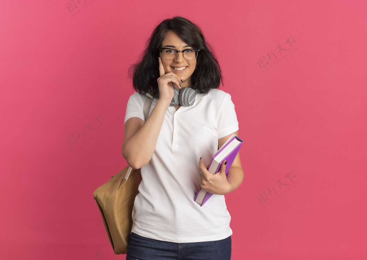 包年轻笑容可掬的白人女学生戴着眼镜 背着书包 手放在脸上 手里拿着粉红色的书 还有复印空间抱粉色女学生