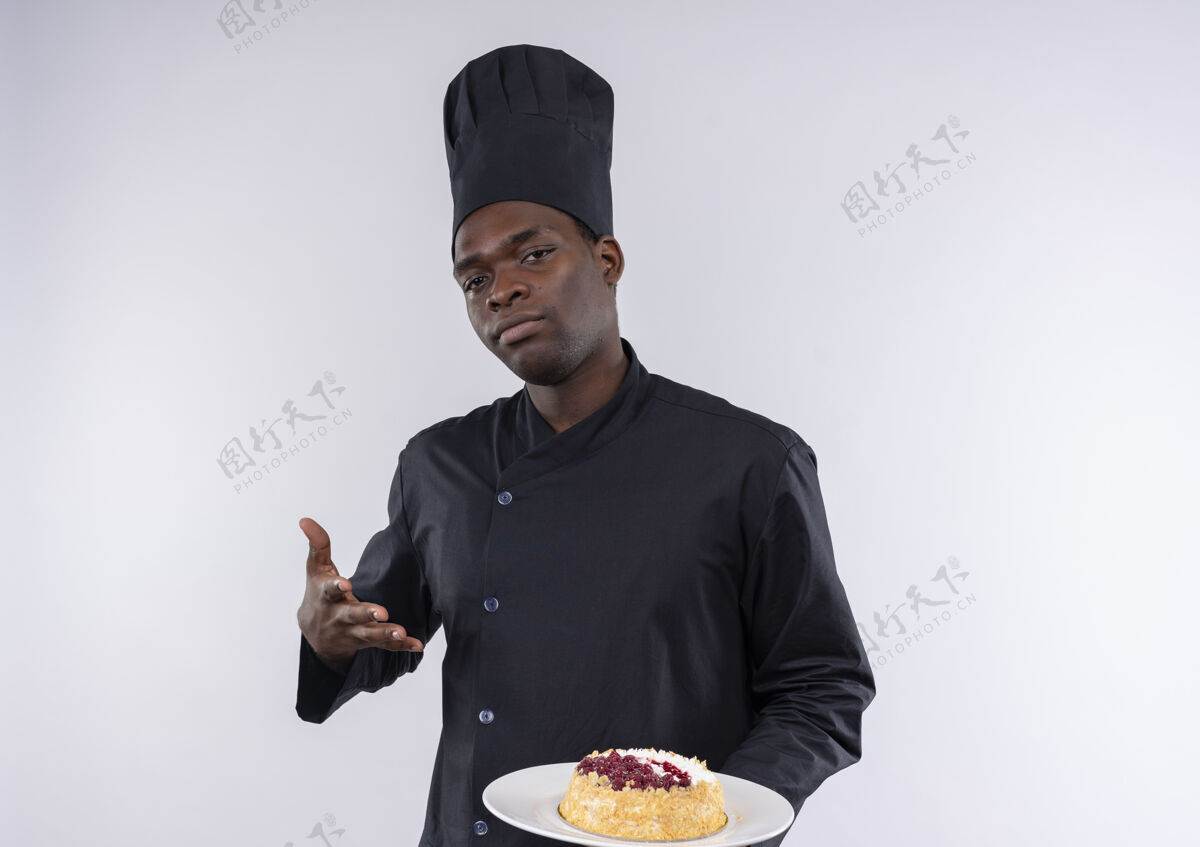 制服身着厨师制服的年轻自信的美国黑人厨师手持并指着盘子上的蛋糕 看着白色的相机 留有复印空间信心蛋糕厨师