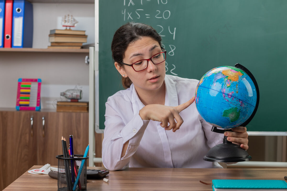 眼镜年轻的女老师戴着眼镜拿着地球仪在教室里讲课 自信地坐在黑板前的课桌旁自信年轻人坐着