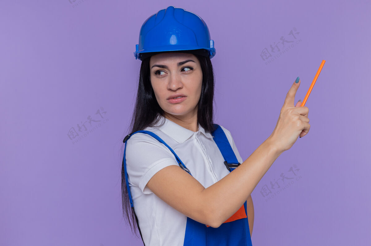 安全身穿建筑制服 头戴安全帽的年轻建筑妇女拿着铅笔指着紫色墙壁上的东西建筑建筑工人什么的