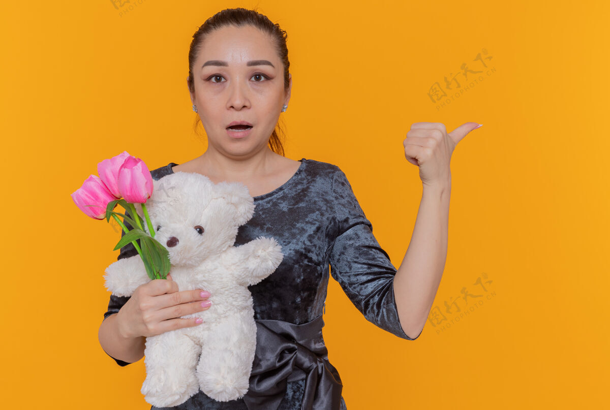 花束困惑的亚洲女人拿着一束粉红色的郁金香和泰迪熊 站在橘色的墙上 用大拇指指着背 看着前方庆祝国际妇女节困惑立场熊