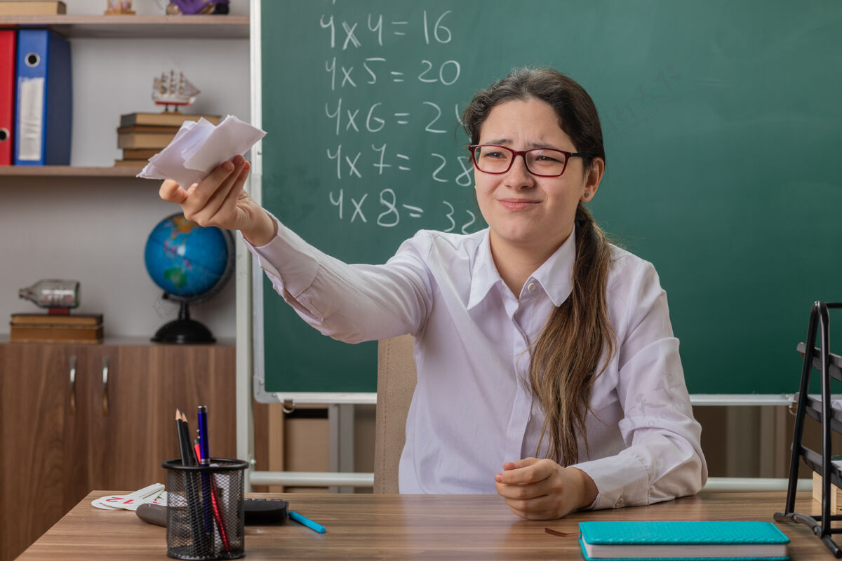 前面年轻的女老师戴着眼镜 手里拿着几张纸 神情不悦地坐在教室黑板前的课桌旁黑板老师年轻人
