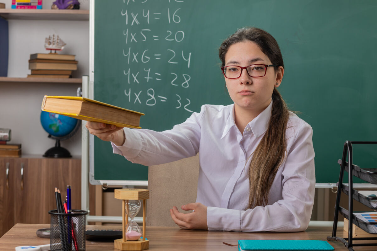 女人年轻的女老师戴着眼镜 在教室的黑板前 自信地坐在课桌旁看书前面年轻人学校