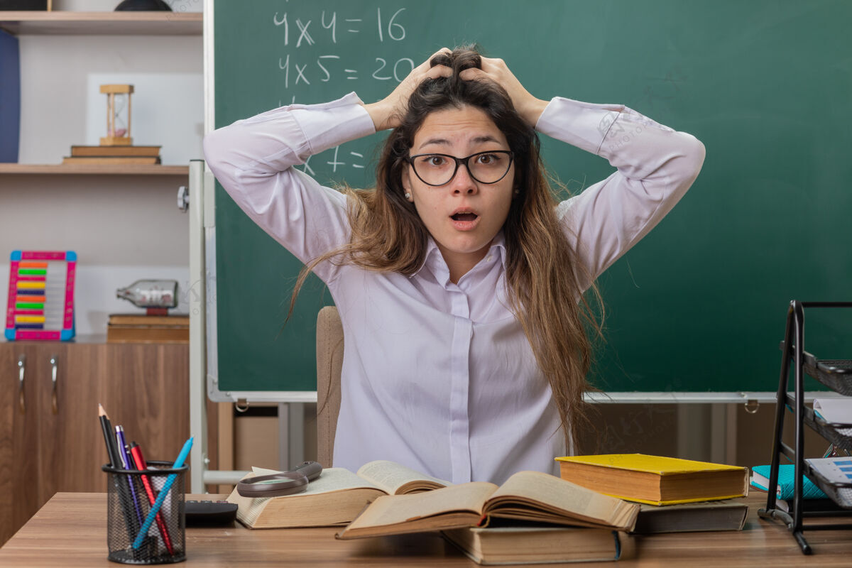 担心年轻的女老师戴着眼镜 拿着书 手放在头上 坐在教室黑板前的课桌旁 神情迷茫 忧心忡忡眼镜困惑教室