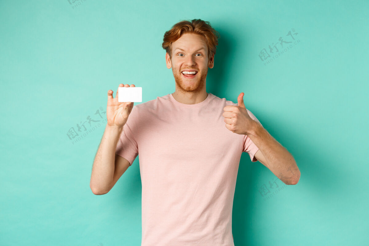 脸兴高采烈的男银行客户穿着t恤衫 竖起大拇指 拿着塑料信用卡 对着镜头满意地微笑 站在绿松石色的背景上成人银行休闲
