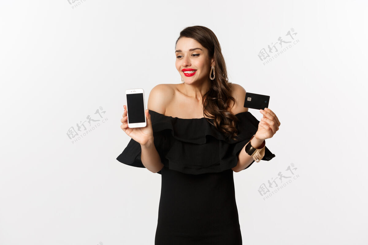 口红时尚和网上购物概念快乐的年轻女子穿着黑色连衣裙 展示信用卡和手机屏幕 站在白色背景上优雅表情女人